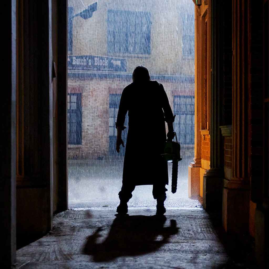 A still from Texas Chainsaw Massacre. Leatherface stands silhouetted in an alleyway with his back to the camera. He holds a chainsaw and gazes out into the rainy street. 