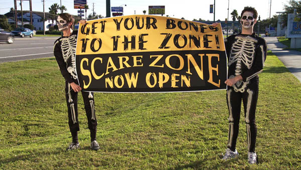 A still from Scare Zone. Two men dressed as skeletons stands by the side of the road holding a sign that says, "Get your bones to the Zone! Scare Zone now open."